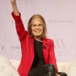 Gloria Steinem speaking warmly at a community gathering surrounded by supporters.