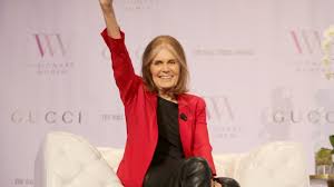 Gloria Steinem speaking warmly at a community gathering surrounded by supporters.